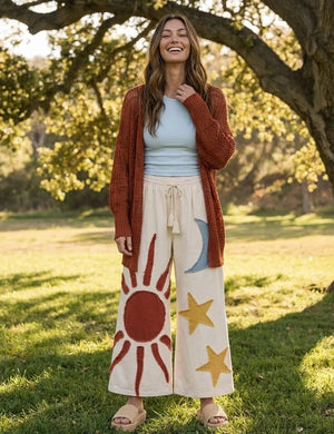 Cream wide leg lounge pants with sun, moon, and star print, styled with rust cardigan and light blue crop top outdoors - 1