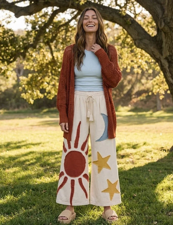 Cream wide leg lounge pants with sun, moon, and star print, styled with rust cardigan and light blue crop top outdoors - 1